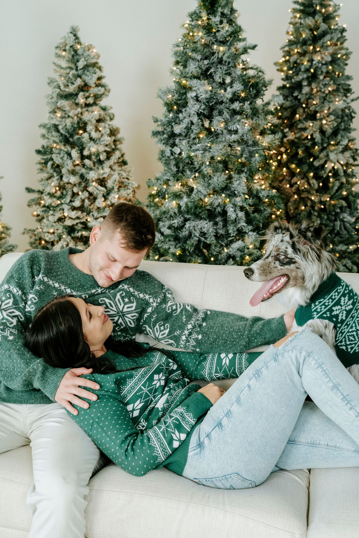 Couple in matching winter sweaters cuddle on white couch with decorated Christmas trees in background.