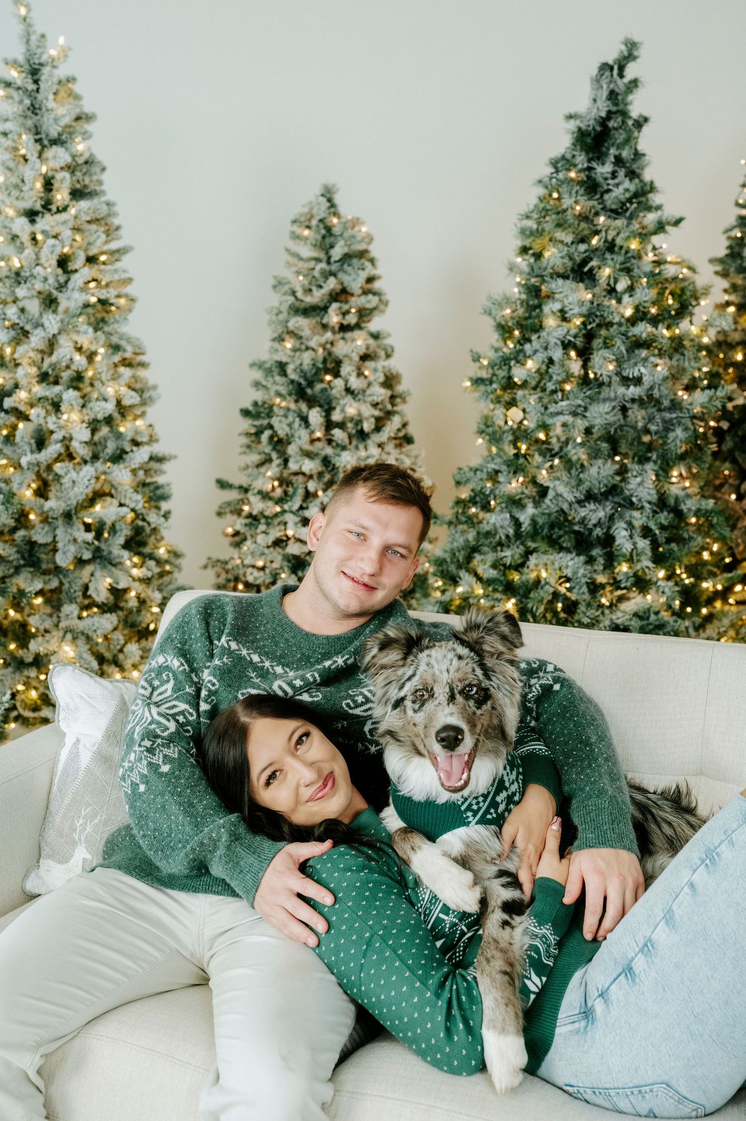 A couple in matching green holiday sweaters cuddles on a white couch with their happy dog against lit Christmas trees.
