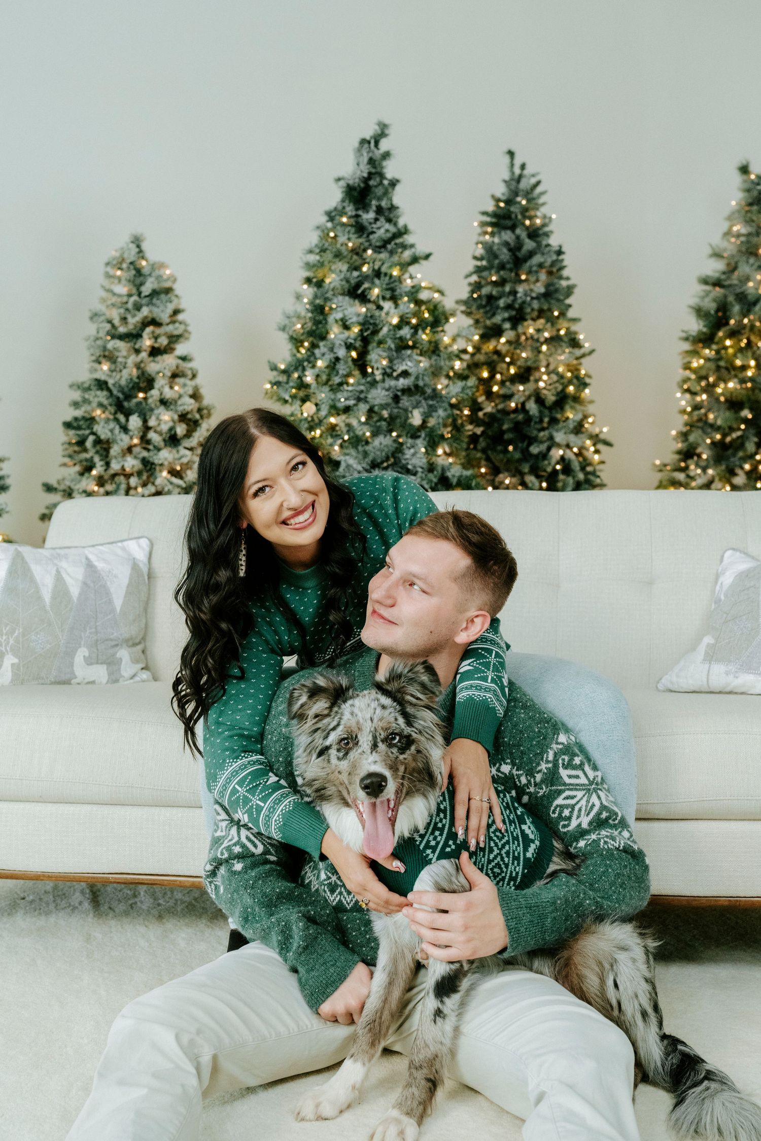 A couple in matching green sweaters sits on a white couch with their dog while decorated Christmas trees glow in background.