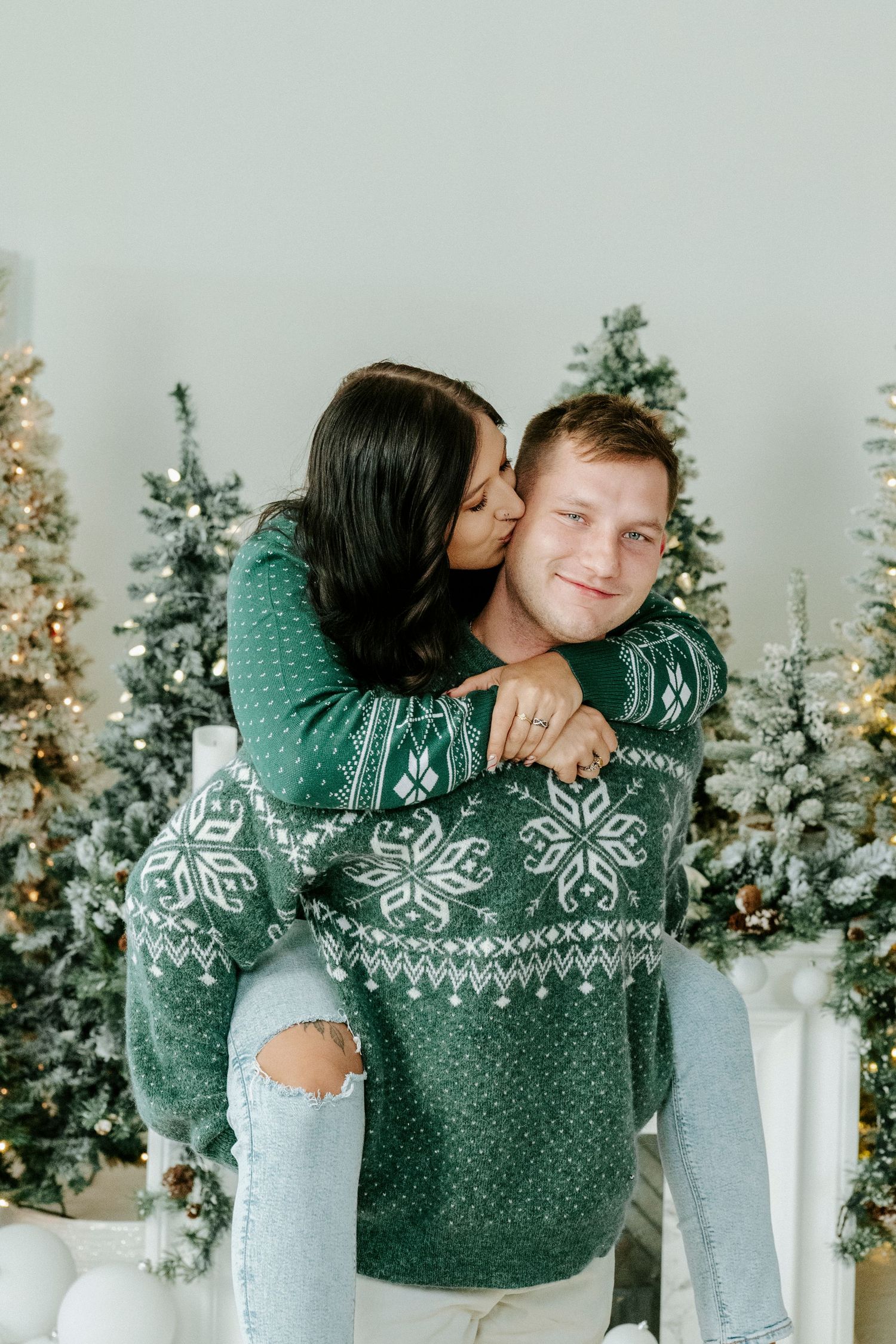 A couple wearing matching winter sweaters embraces in front of decorated Christmas trees and white pillars.
