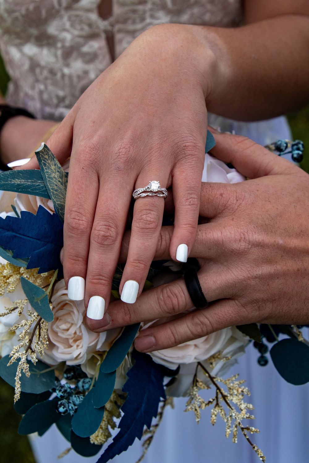 Close-up of hands showing off engagement rings with white manicured nails and blue and cream floral bouquet details.
