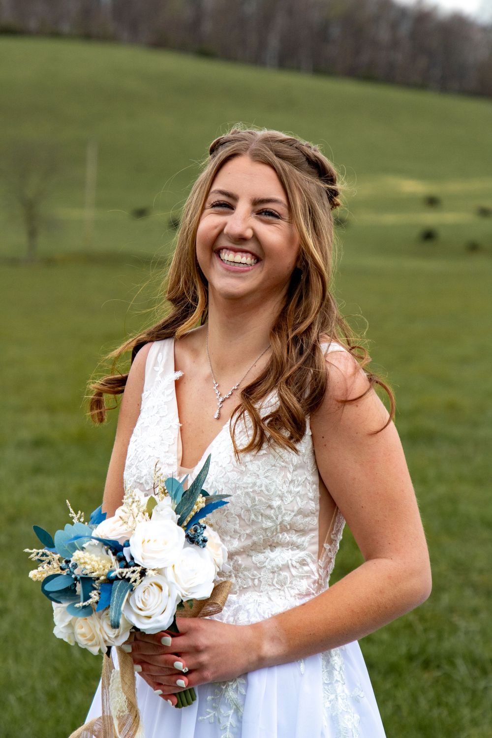 A bride in a white lace dress holds a blue and white floral bouquet while standing in a green field.