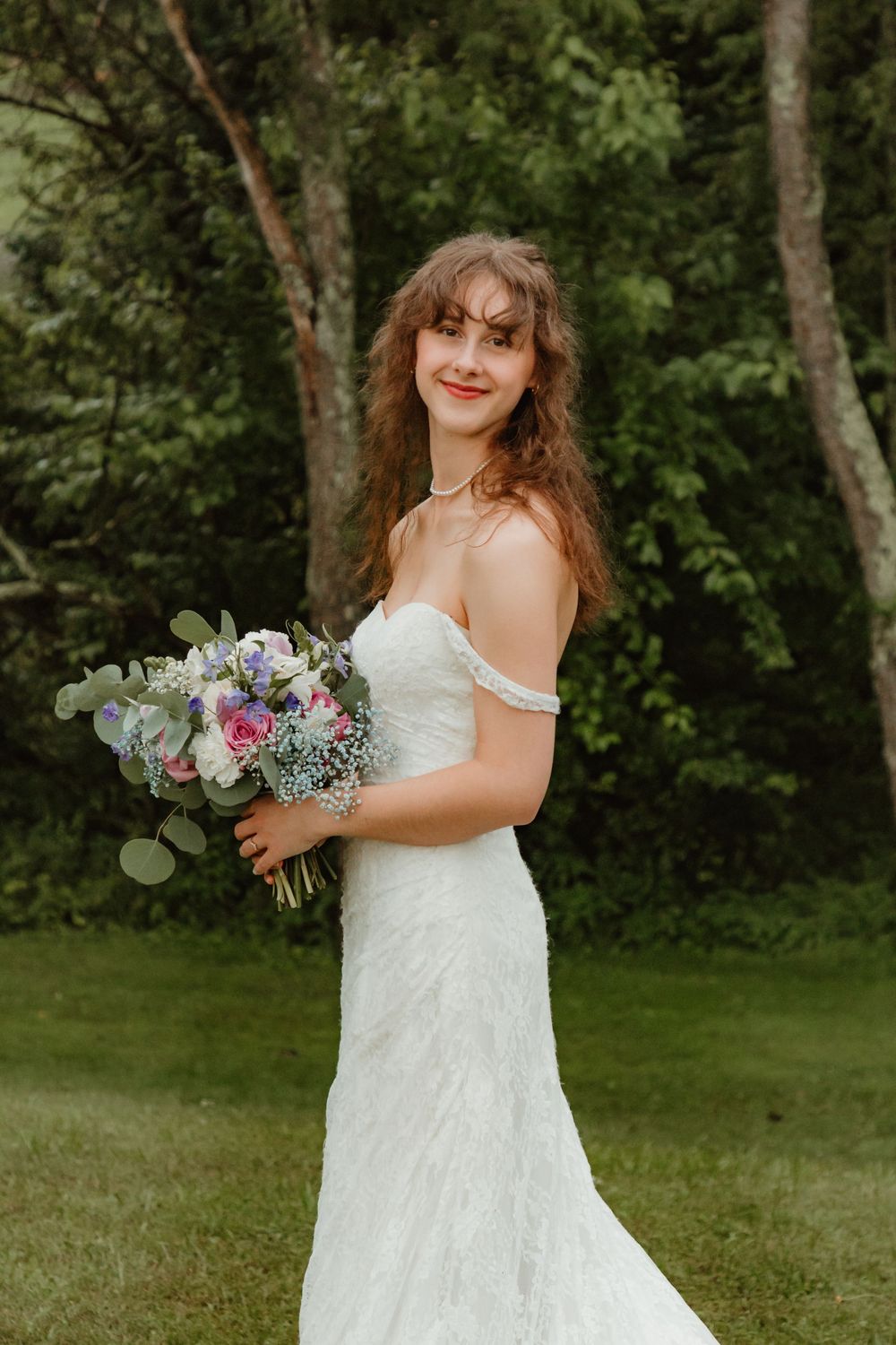 Bride in a white off-shoulder wedding dress holds a pink and purple bouquet while posing in a garden setting.