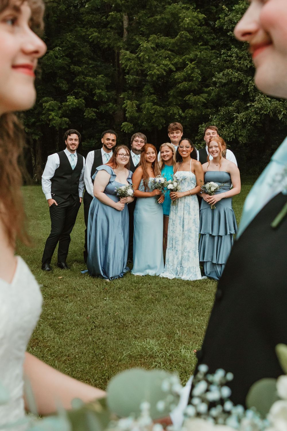 Wedding party in light blue bridesmaid dresses poses together on green lawn during outdoor celebration.
