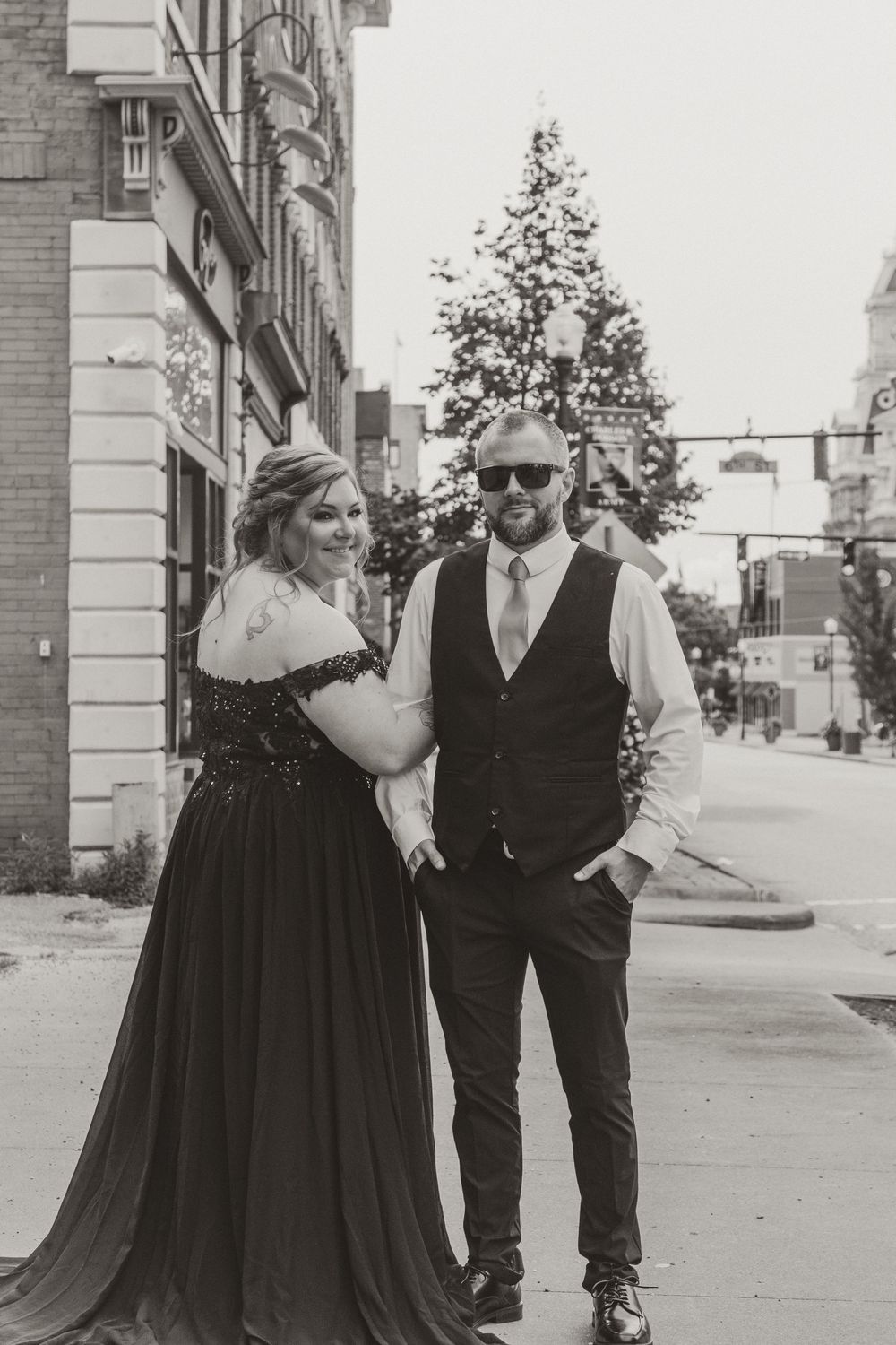 A couple in formal attire poses together on a city street, with the person wearing a black dress and another in a vest.