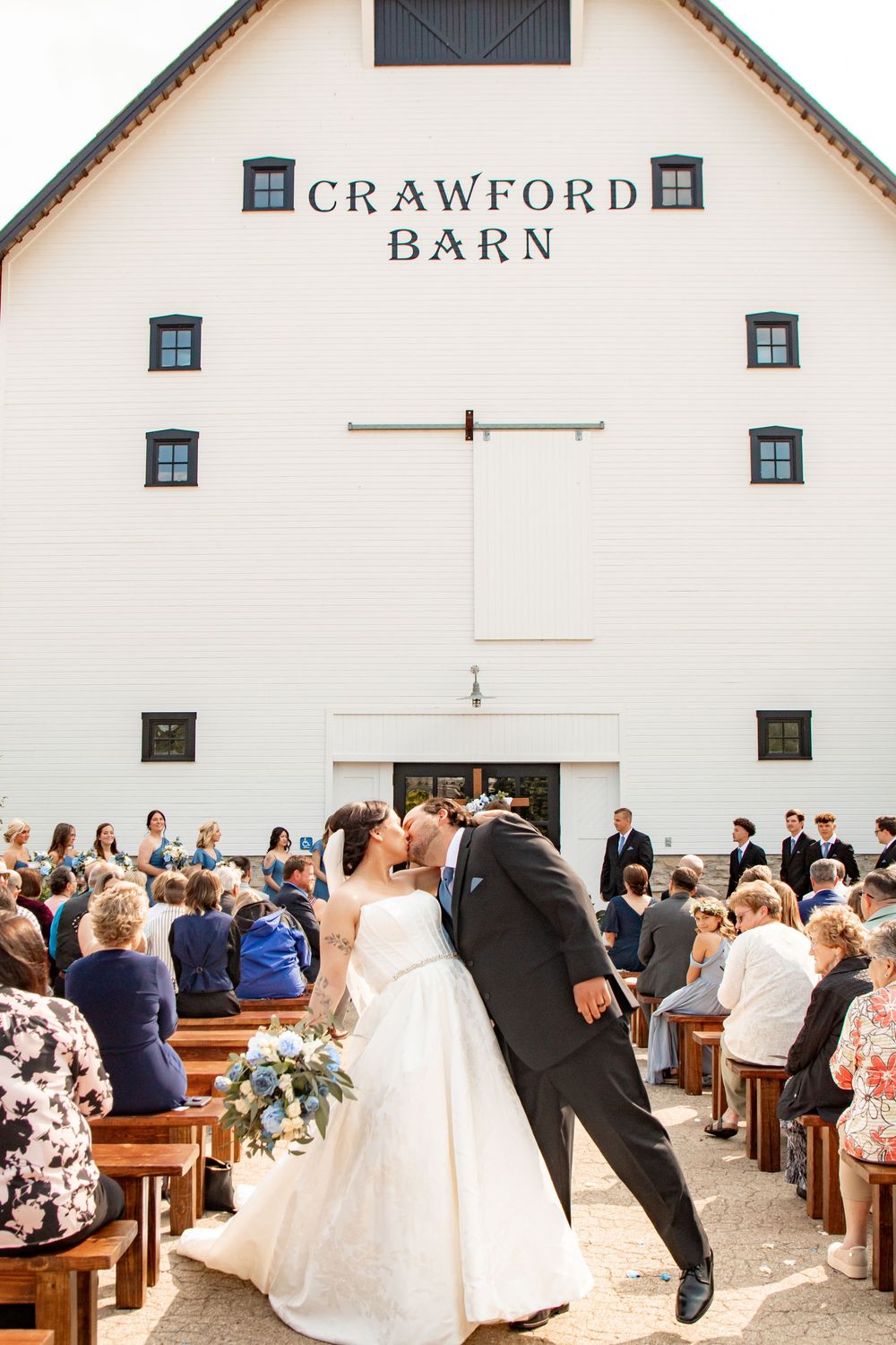 Wedding ceremony taking place inside a rustic white barn venue called Crawford Barn with wooden pew seating.