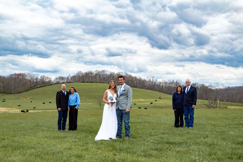 Small wedding ceremony takes place in a rolling pastoral field with scattered guests under dramatic clouds.