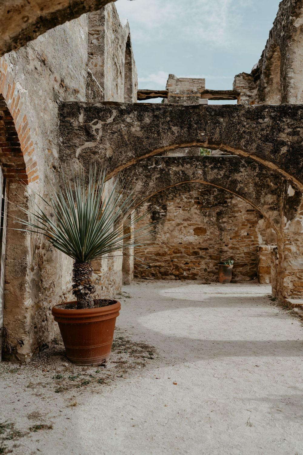 Stone archways and potted plants line a rustic courtyard pathway in warm sunlight.