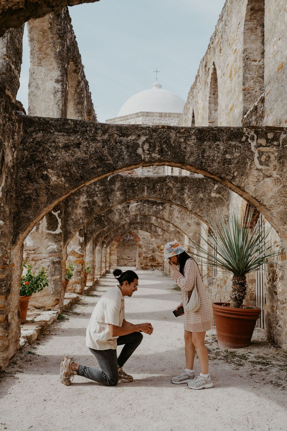 A surprise proposal moment unfolds under stone archways at a historic mission.