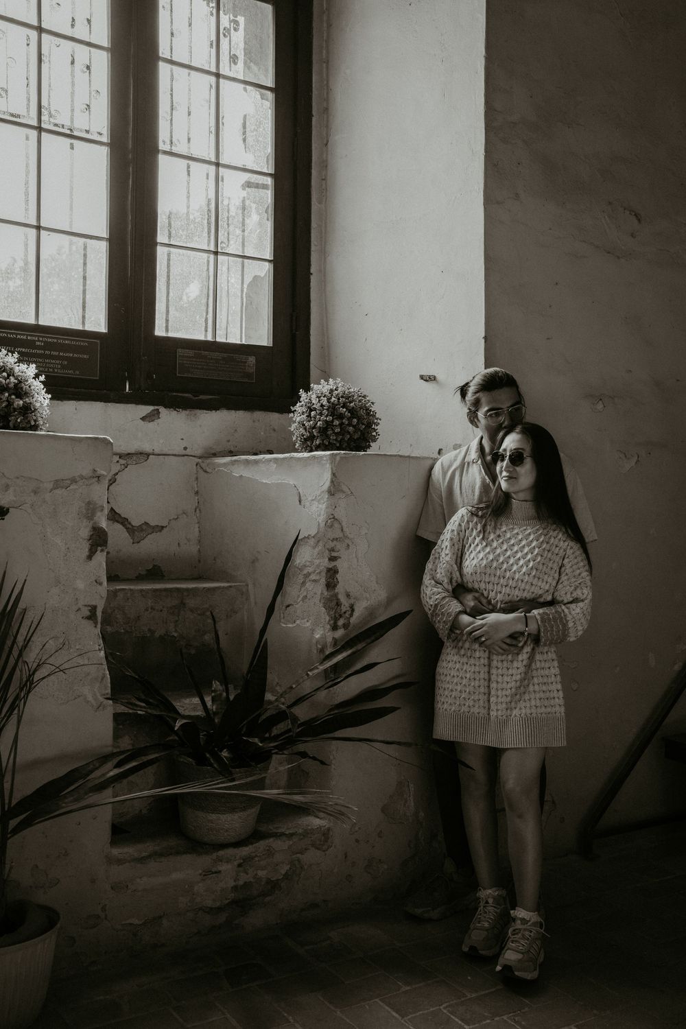 A couple poses by large windows in a vintage-style black and white photo.