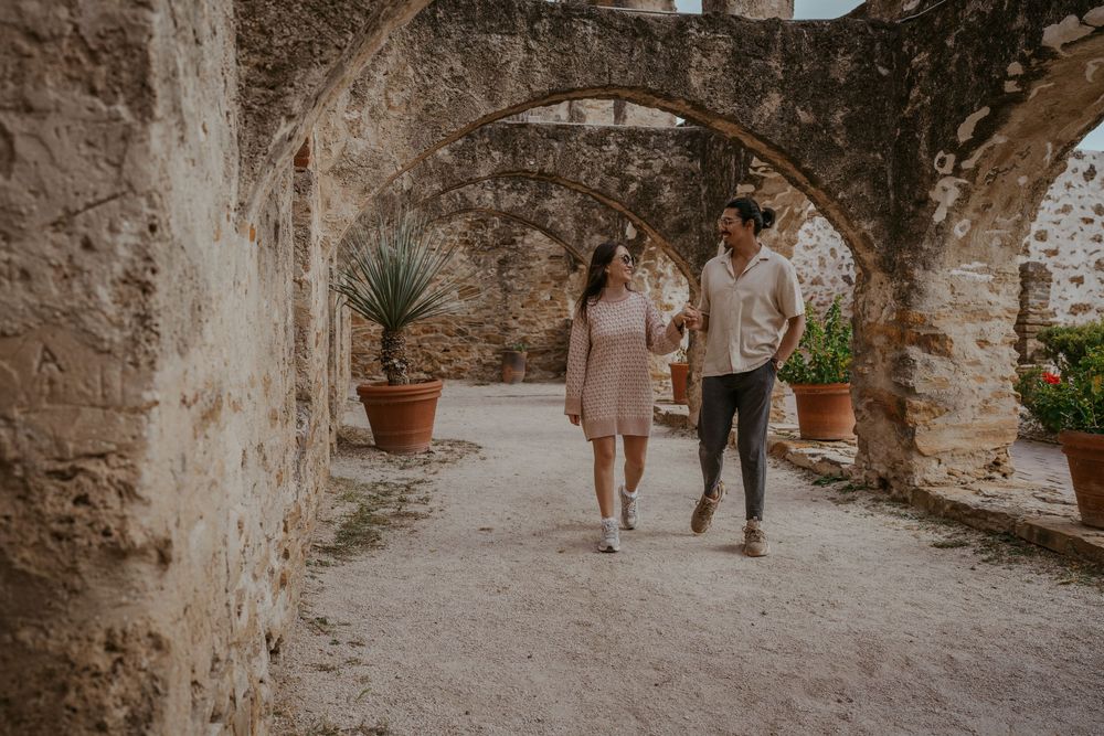 Couple walking along a rustic stone corridor with arched windows.