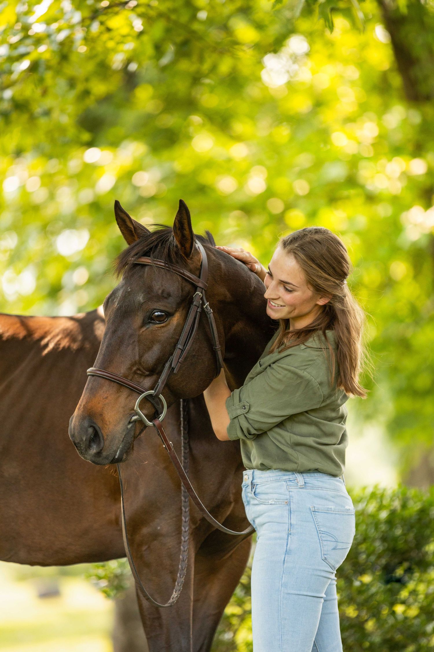 A person in jeans and green shirt shares a tender moment with their dark brown horse in a sunlit outdoor setting.