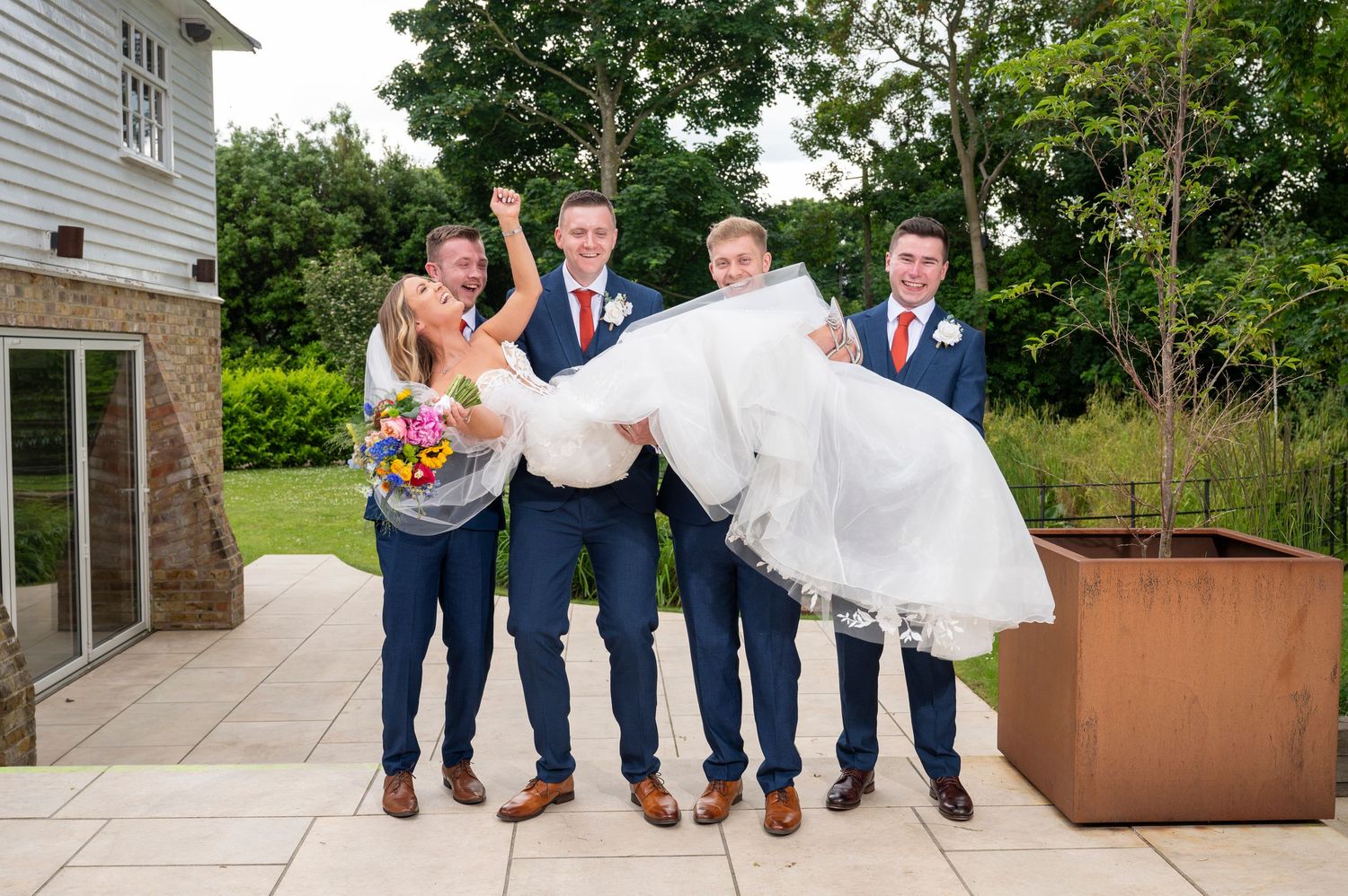 Groomsmen in navy suits and brown shoes lift bride in white dress while posing outdoors for wedding photos.
