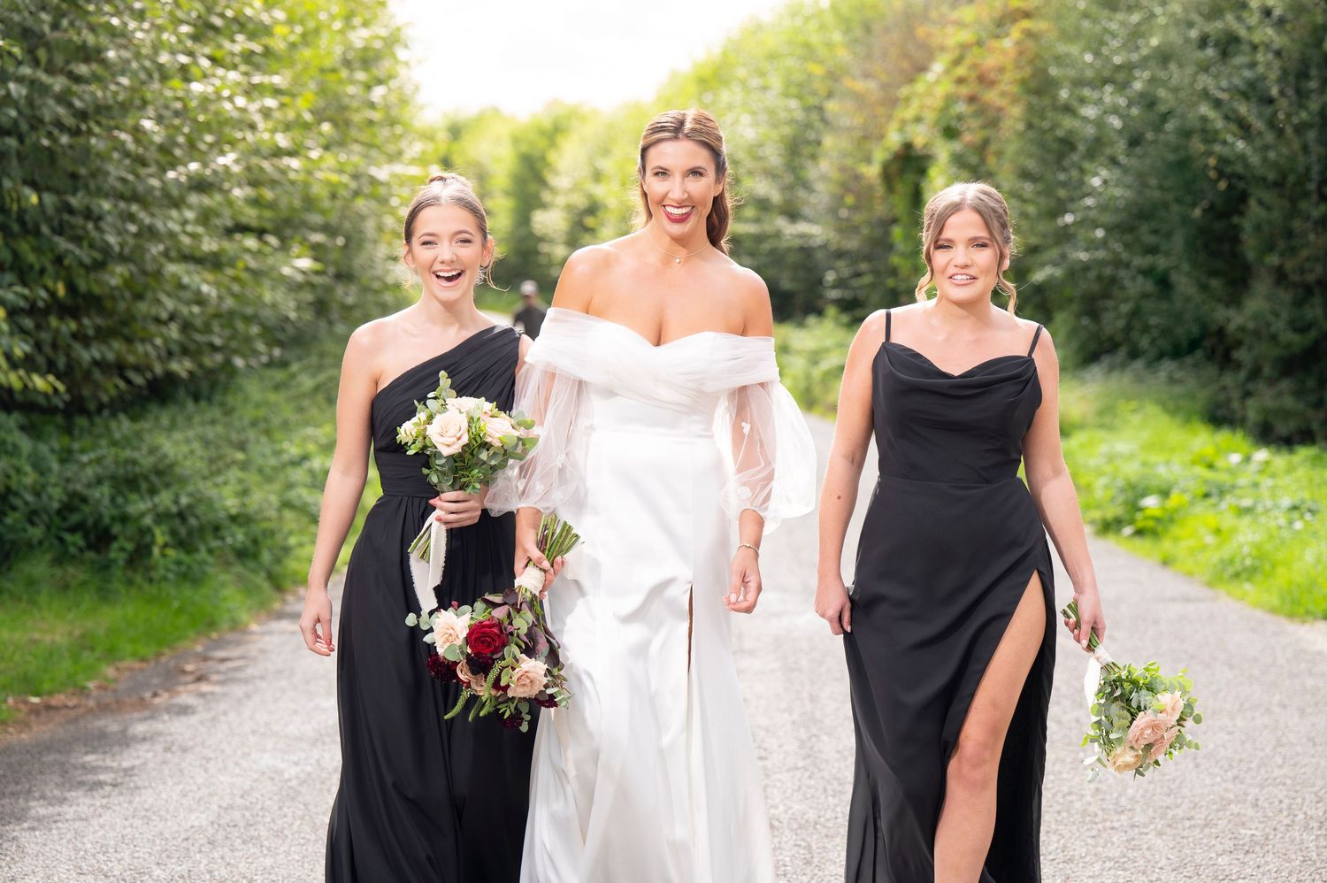 Three women in formal dresses walk together on a country path holding bouquets at an outdoor wedding celebration.