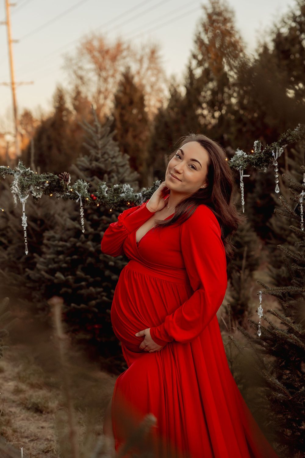 A woman in a flowing red dress poses gracefully among evergreen trees at sunset.
