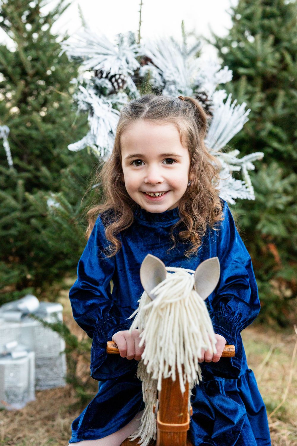 Child in blue velvet dress plays with hobby horse toy against evergreen tree backdrop.