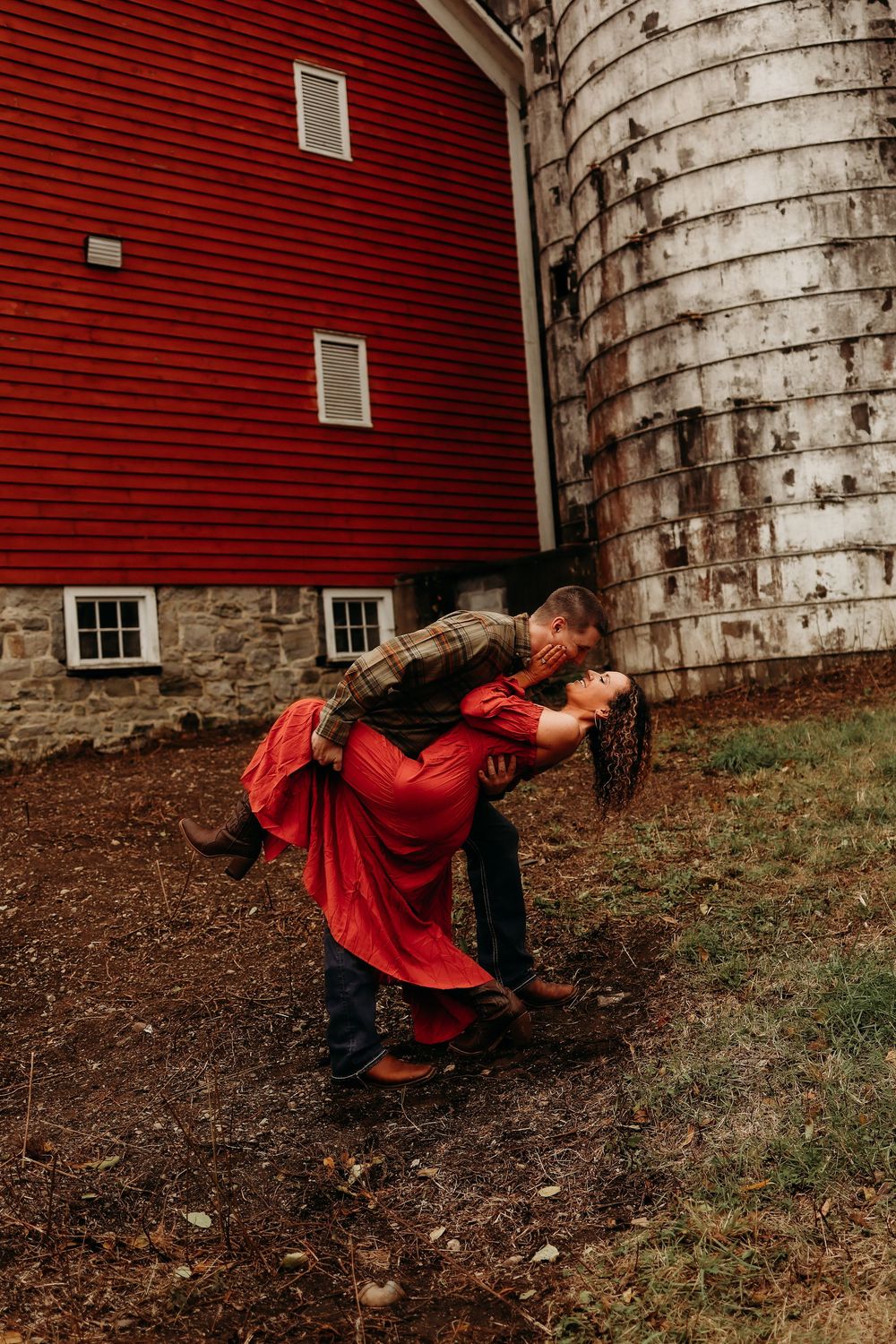 A romantic couple embraces in front of a rustic red barn and stone silo on a rural farm property.