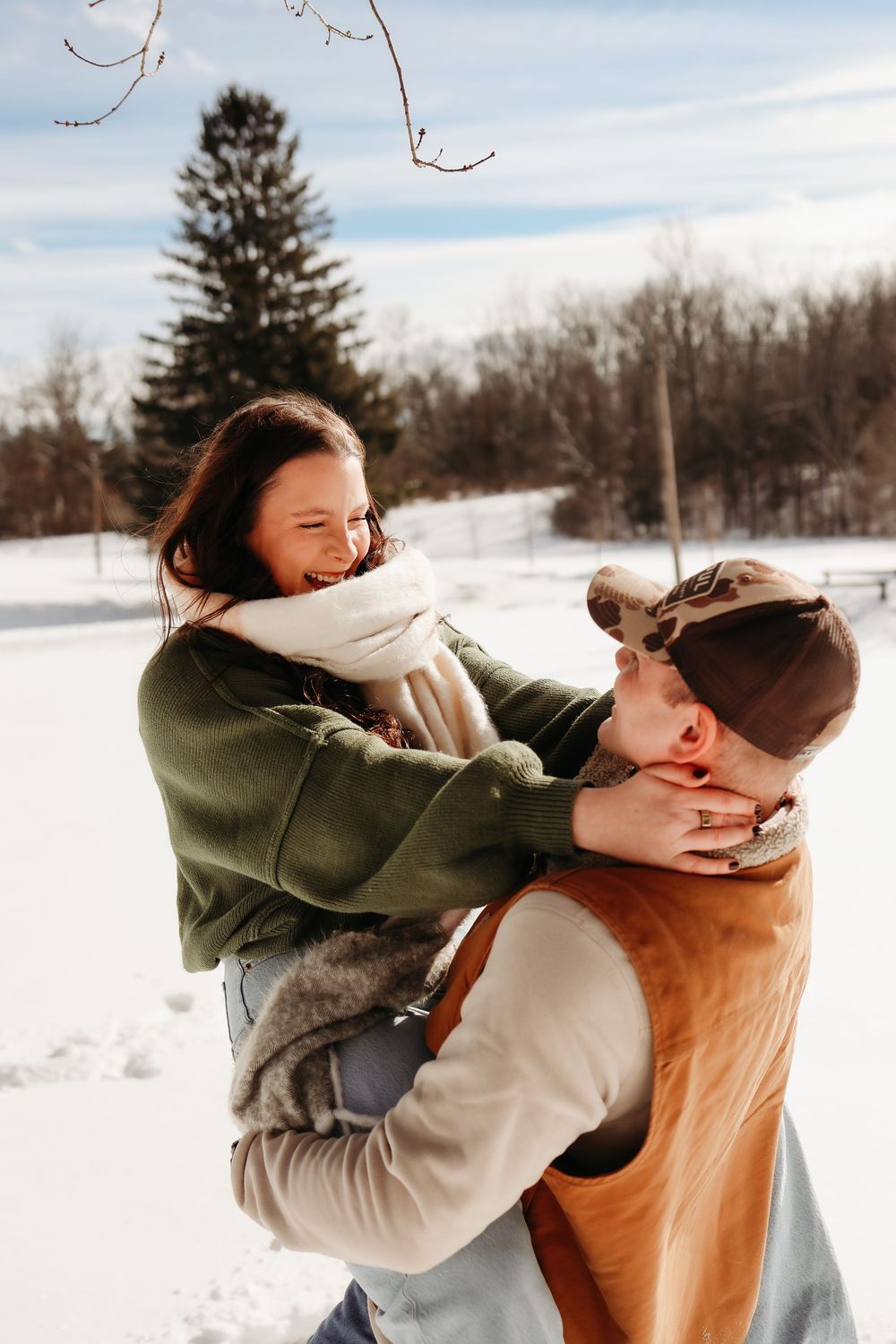A couple shares a joyful embrace in a snowy winter landscape with evergreen trees in the background.
