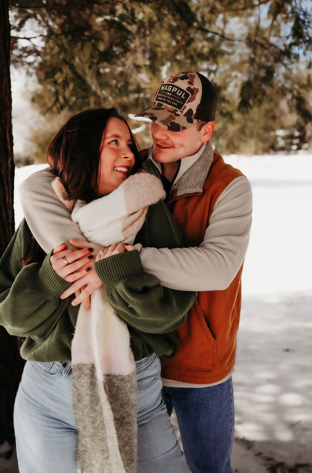A couple embraces playfully in a snowy winter setting wearing casual sweaters and jeans.