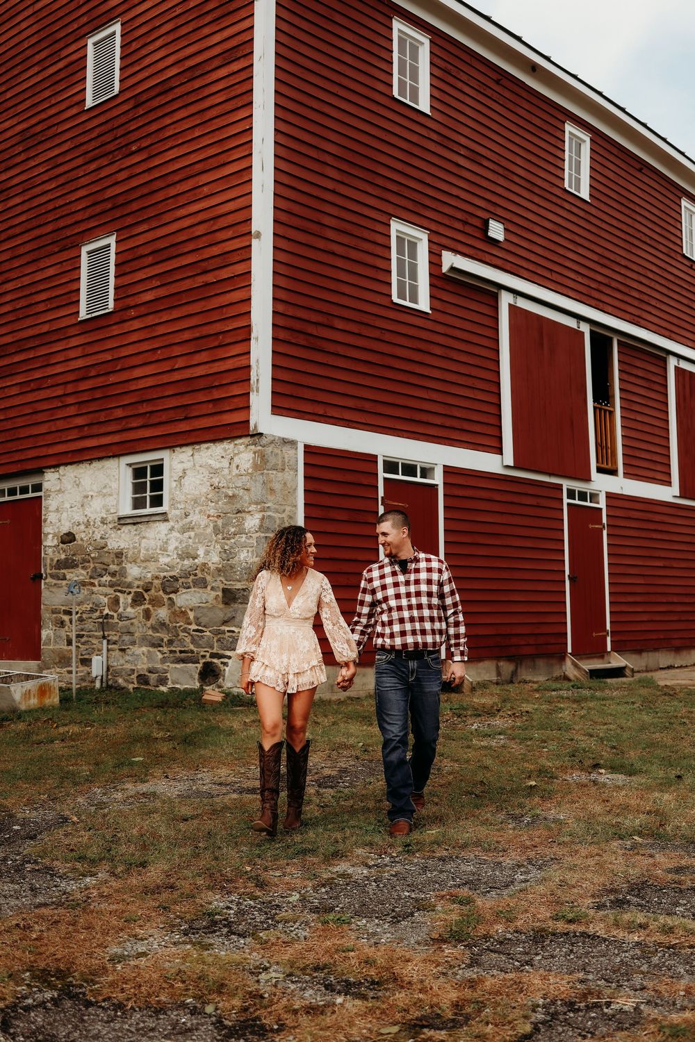A couple holds hands while walking beside a classic red barn with stone foundation on a farm.