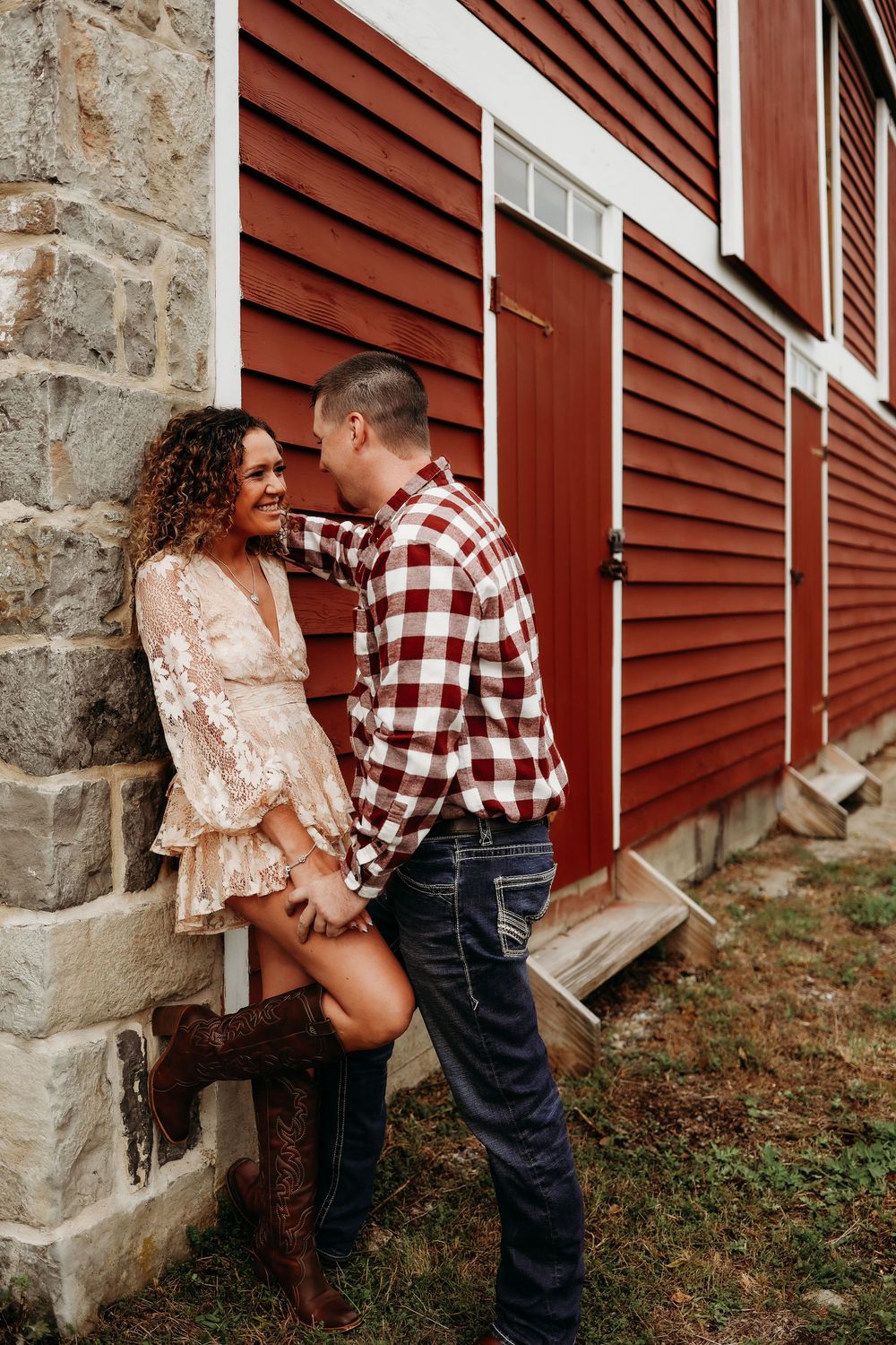 A couple in rustic attire poses together against a red barn wall with stone foundation during a countryside photoshoot.