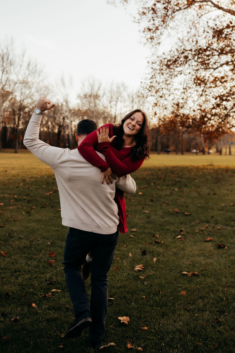 Two people share playful moments in an autumn field with fallen leaves and bare trees in the background.