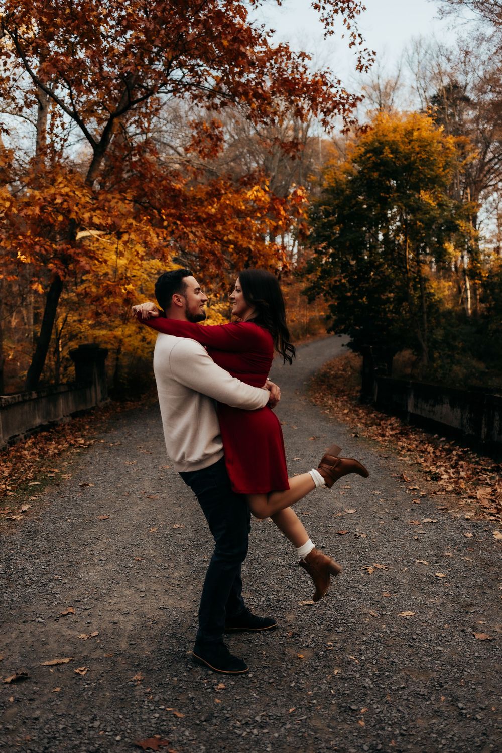 A couple dances playfully on a leaf-covered path surrounded by autumn trees with vibrant orange and red foliage.