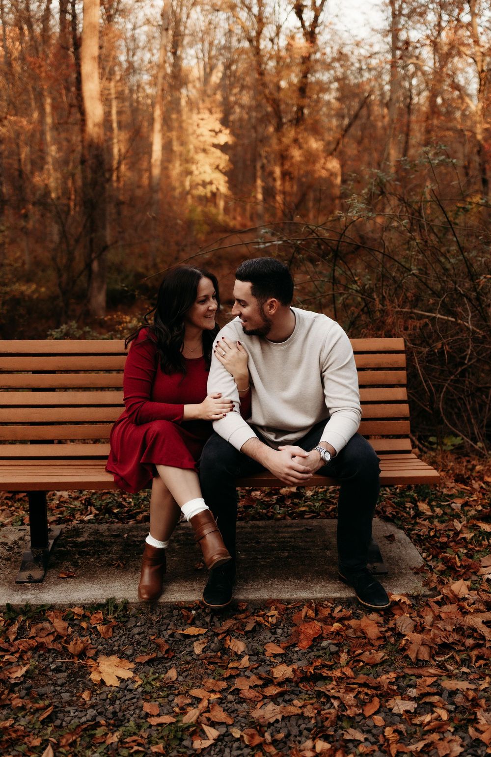 Couple sitting together on wooden bench in autumn forest with fallen leaves and warm lighting.