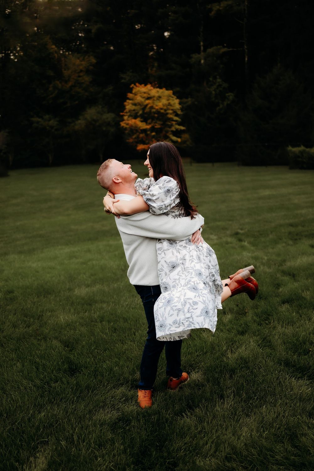 A couple embrace in a joyful moment on grass with autumn trees in the background.