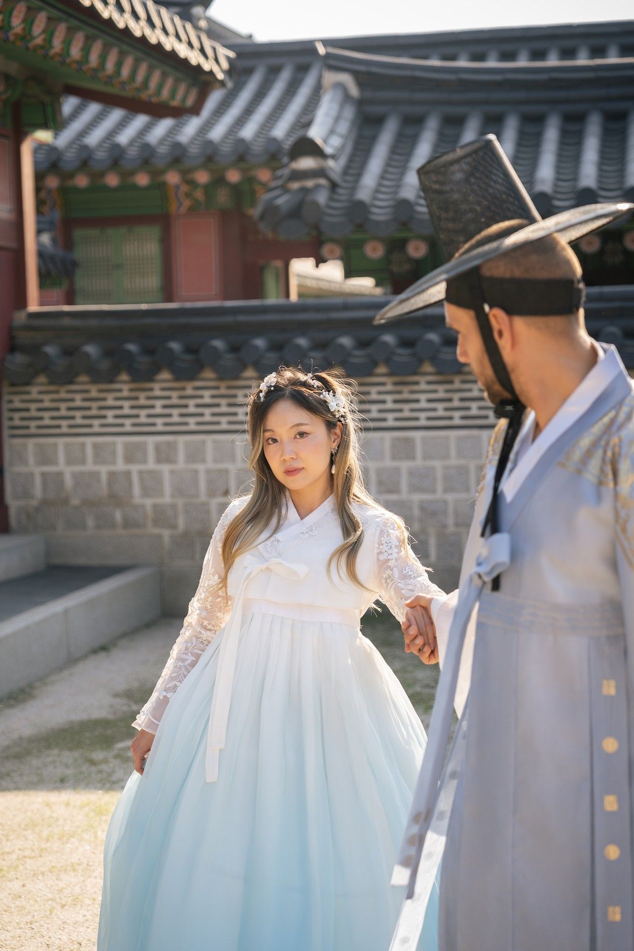 A couple in traditional Korean hanbok attire walking through a palace courtyard with traditional architecture.