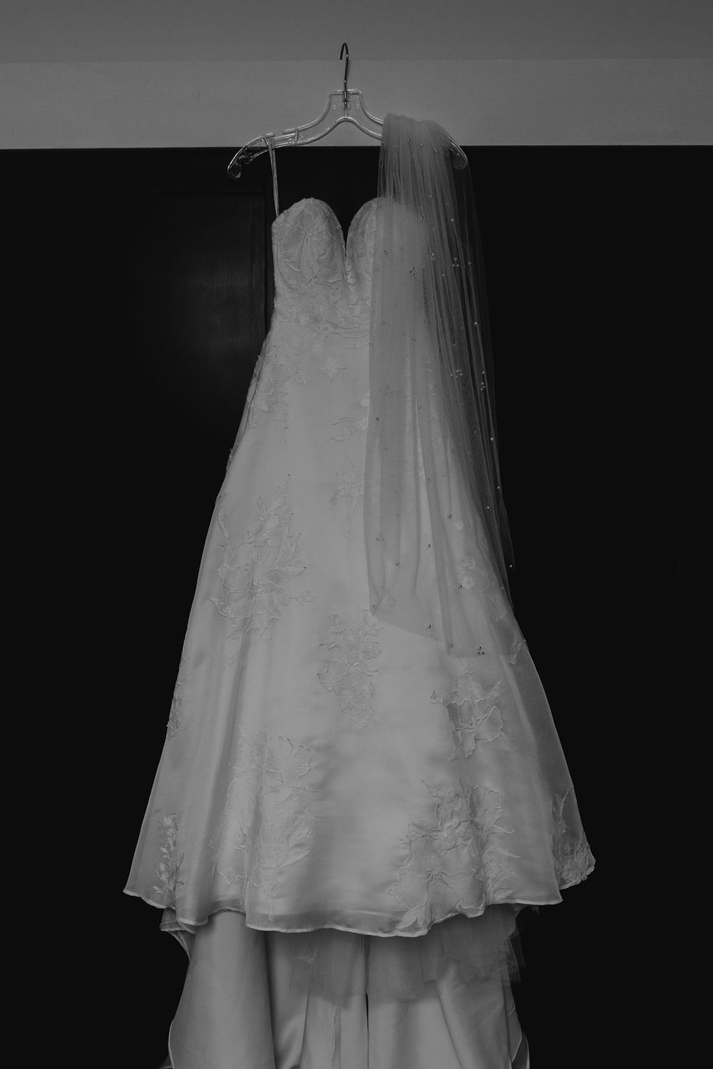 Wedding dress hanging against dark background showing intricate details and flowing layers.