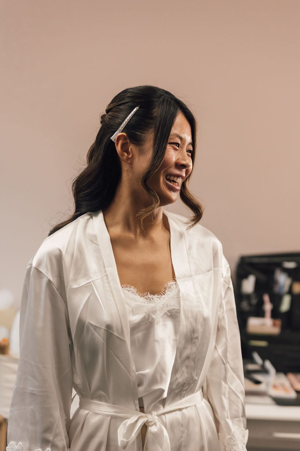 Bride wearing white silk robe smiles during wedding day preparations.