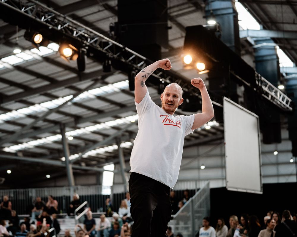 Performers execute dynamic dance moves in an indoor venue with stage lighting and audience members visible.