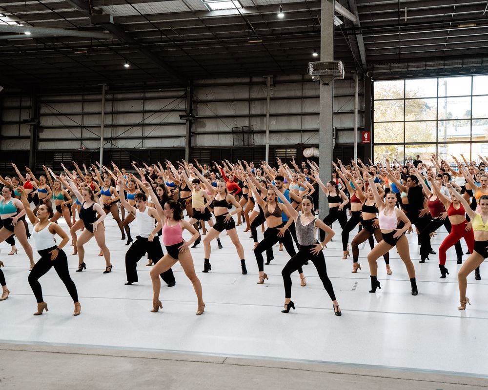 Large group of dancers practicing synchronized choreography in a spacious industrial warehouse studio space.