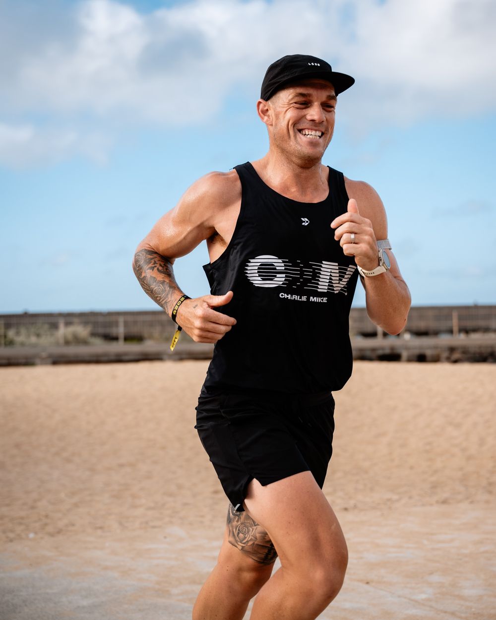 Athletic figure in black tank top and shorts jumps playfully on a sandy beach with cloudy sky in background.
