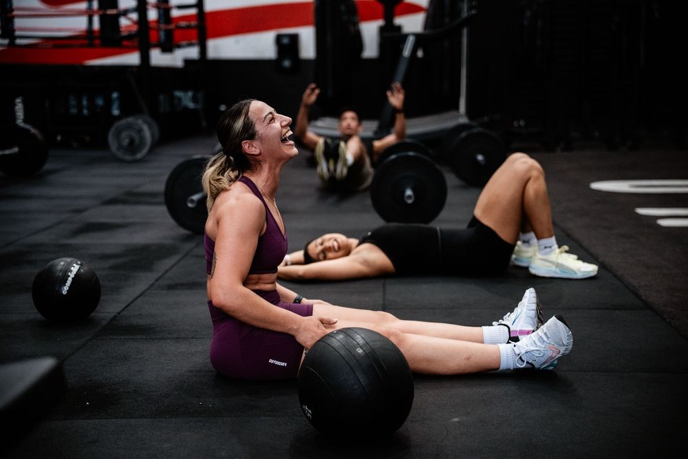 Exhausted athletes lie on gym floor after intense CrossFit workout with medicine balls and weights.