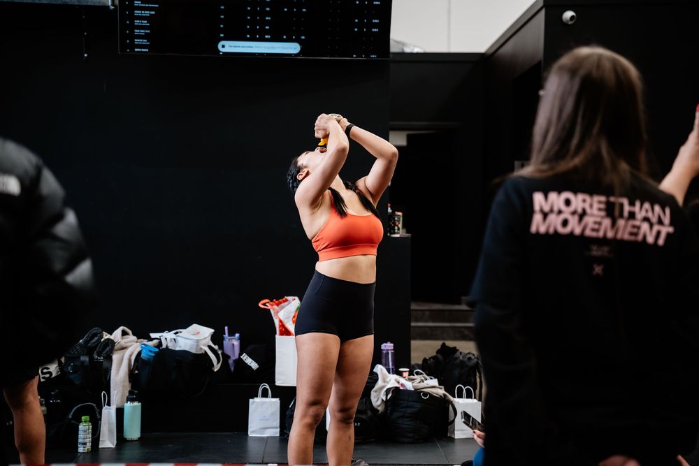 Athlete in orange sports bra and black shorts performs overhead barbell lift in dark gym setting.