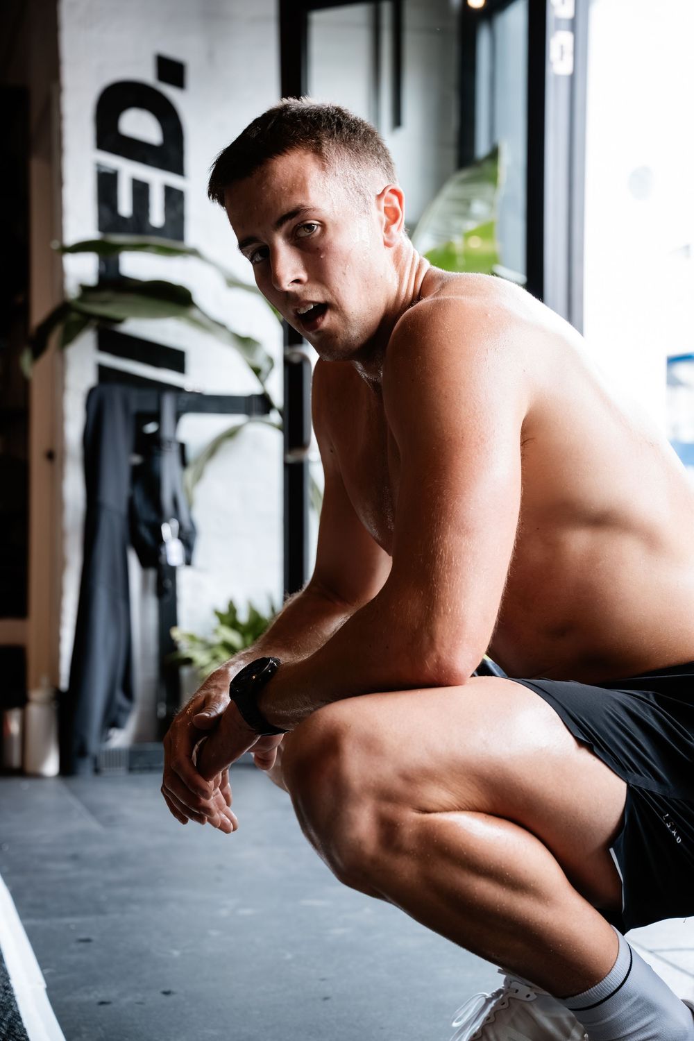 Shirtless athlete performs intense bodyweight exercises on a gym floor near large windows.