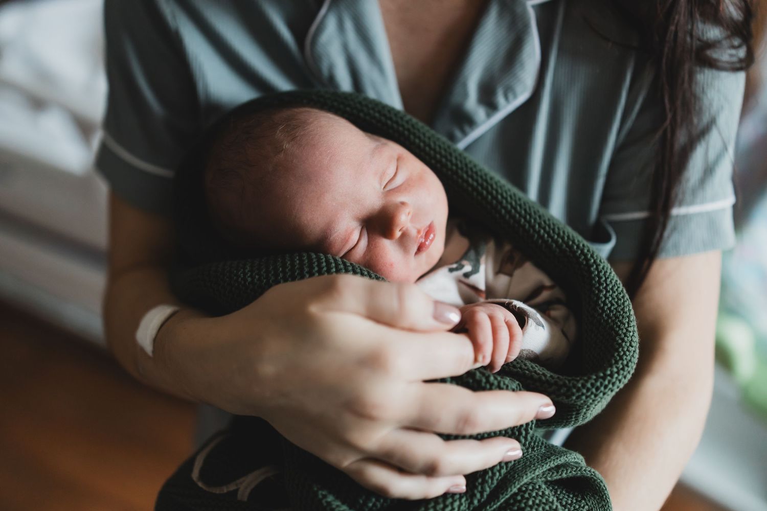 A newborn baby wrapped in a green knit blanket sleeps peacefully in someone's arms.