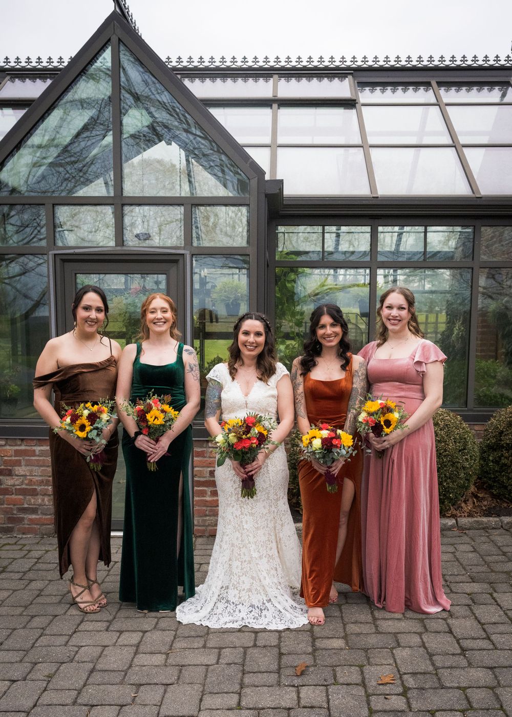 Bridal party poses with colorful sunflower bouquets in front of glass greenhouse.