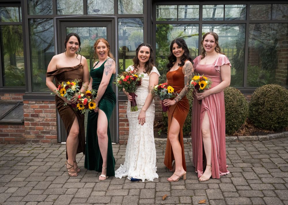 Wedding party in contrasting dresses holding vibrant sunflower bouquets pose together on brick patio.