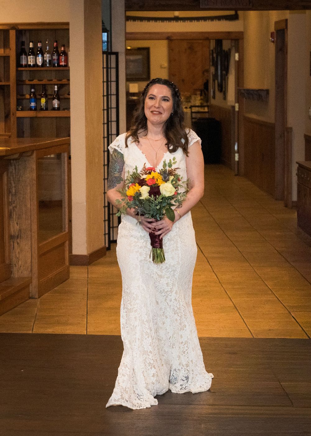 A bride in a lace wedding dress holds a colorful bouquet while standing in a venue hallway.