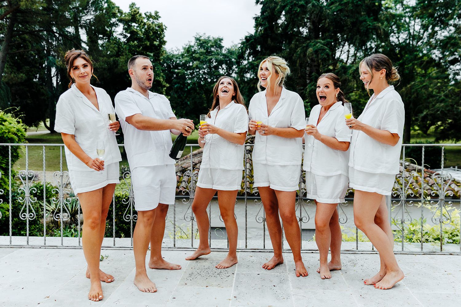 A group of friends in matching white outfits stand together on a balcony surrounded by greenery.