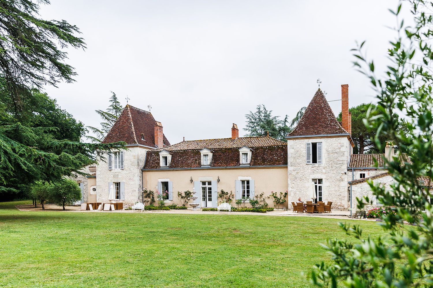 Elegant French chateau with white stone walls and red tiled turrets sits on manicured lawn surrounded by trees.