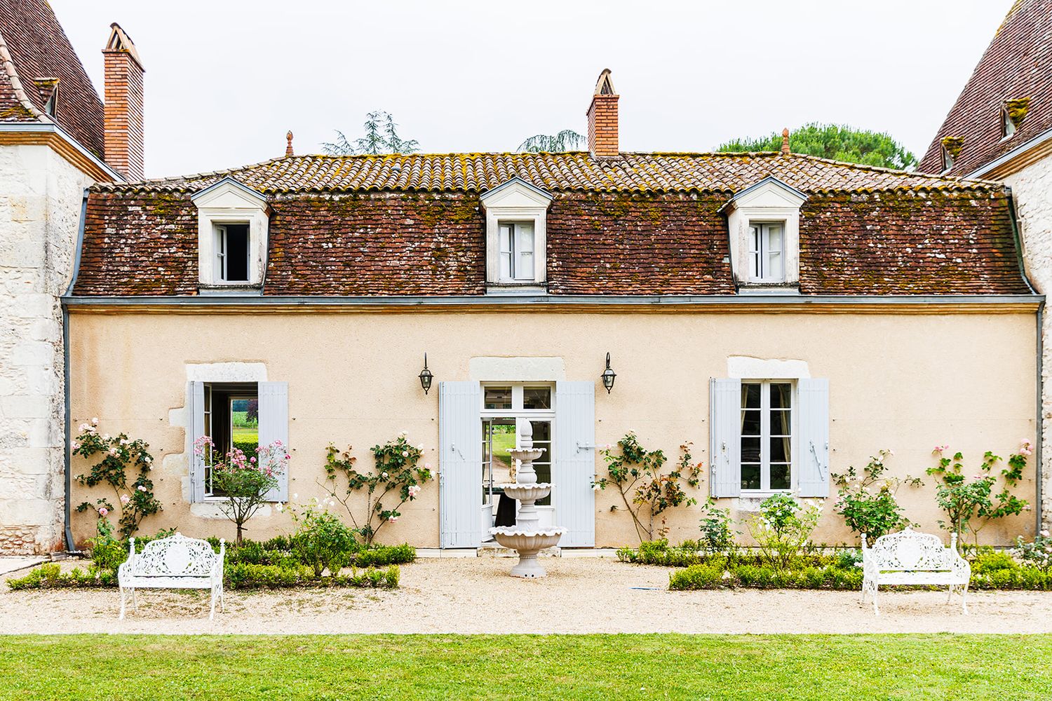 Charming French country house with white walls, dormer windows, climbing vines and garden chairs on the lawn.