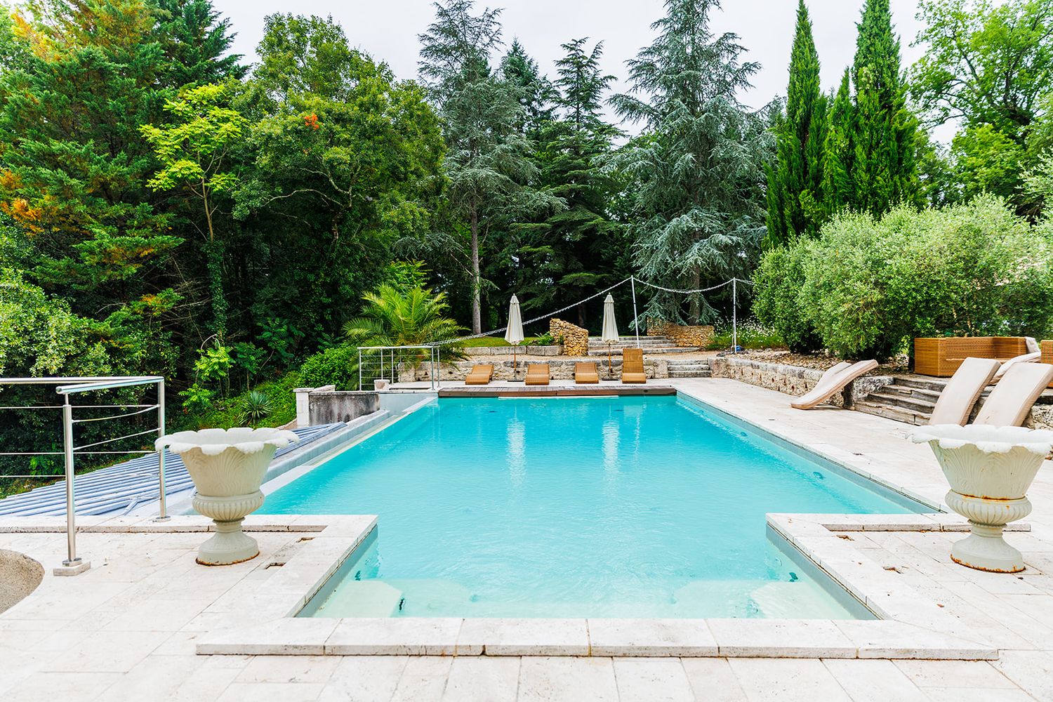 Luxurious turquoise swimming pool surrounded by lush green trees and white lounge chairs in an elegant garden setting.