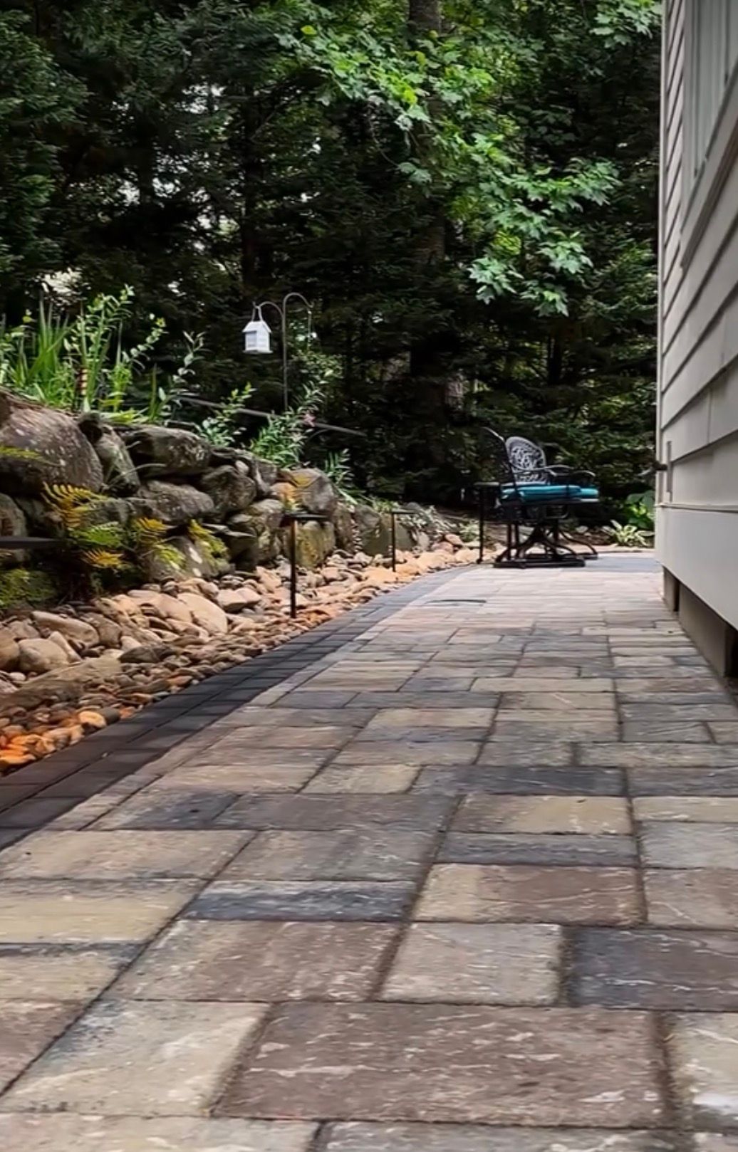 Stone paver patio with landscaped rock border extends along the side of a house in a wooded setting.