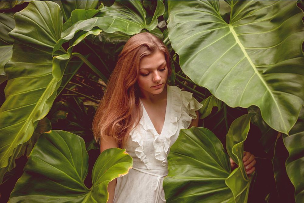 A woman in a white dress poses among large tropical elephant ear leaves in a lush garden setting. Photo session.