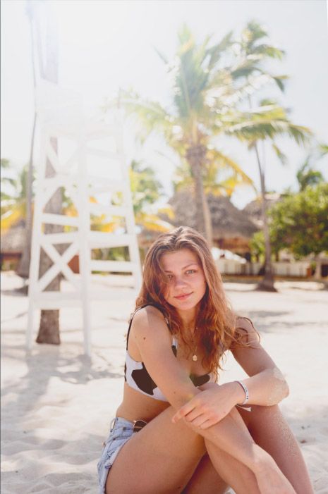 A woman in casual attire sits on a sandy beach with palm trees and white structures in the tropical background. Photo session.