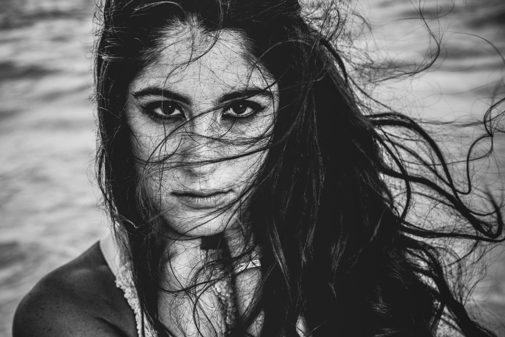 A dramatic black and white portrait shows windswept dark hair framing a face with intense eyes gazing at the camera. Cap Cana Juanillo
